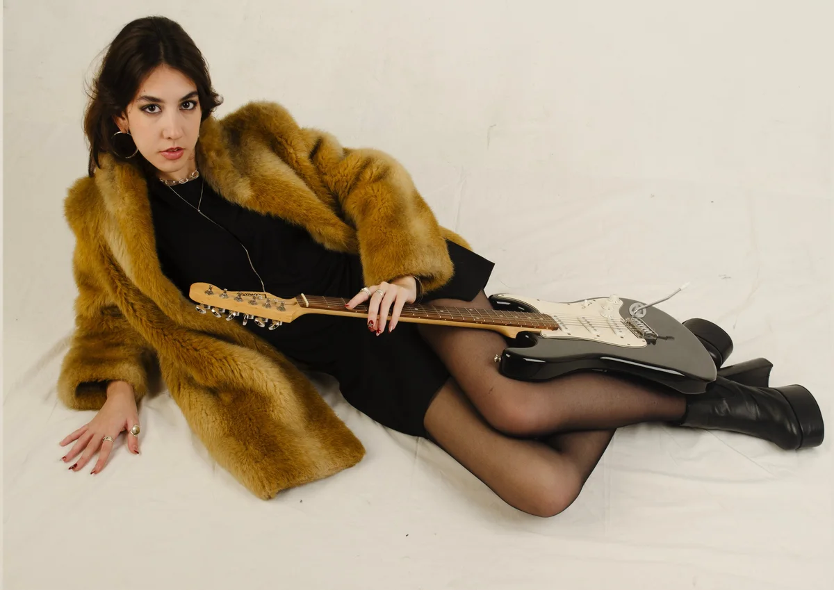 Modelo recostada con tapado de piel y guitarra eléctrica sobre fondo blanco
