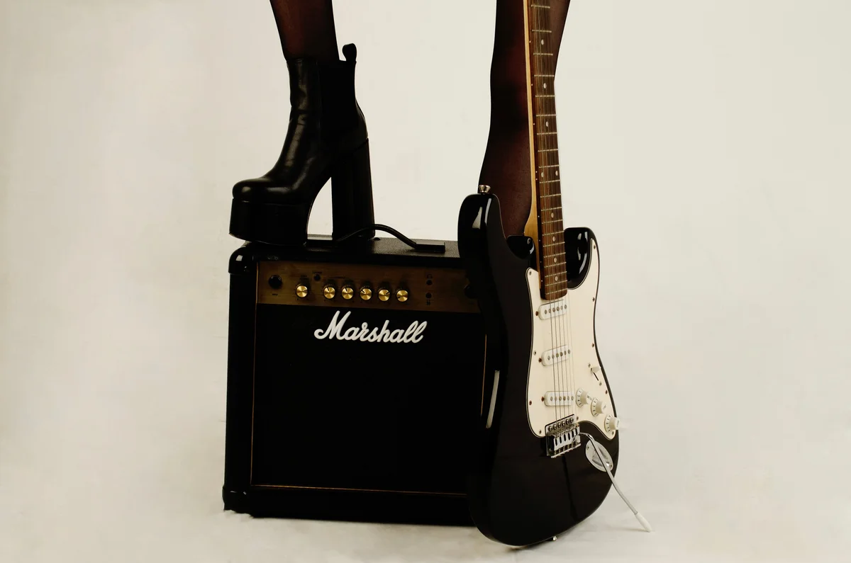 Piernas con medias y botas de plataforma junto a un amplificador Marshall y una guitarra