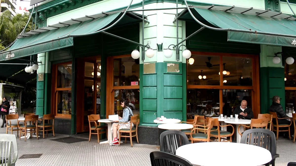 Fotograma del fashion film: fachada verde de un café de esquina con mesas en la vereda
