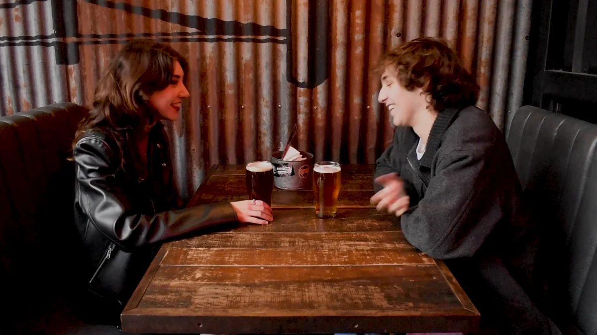 Fotograma del fashion film: dos jóvenes conversan en la mesa de madera de un bar con paredes de chapa