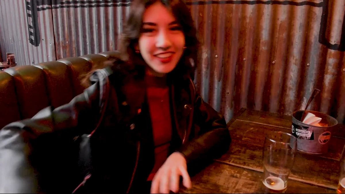 Fotograma del fashion film: una joven con campera de cuero sonríe sentada en un bar